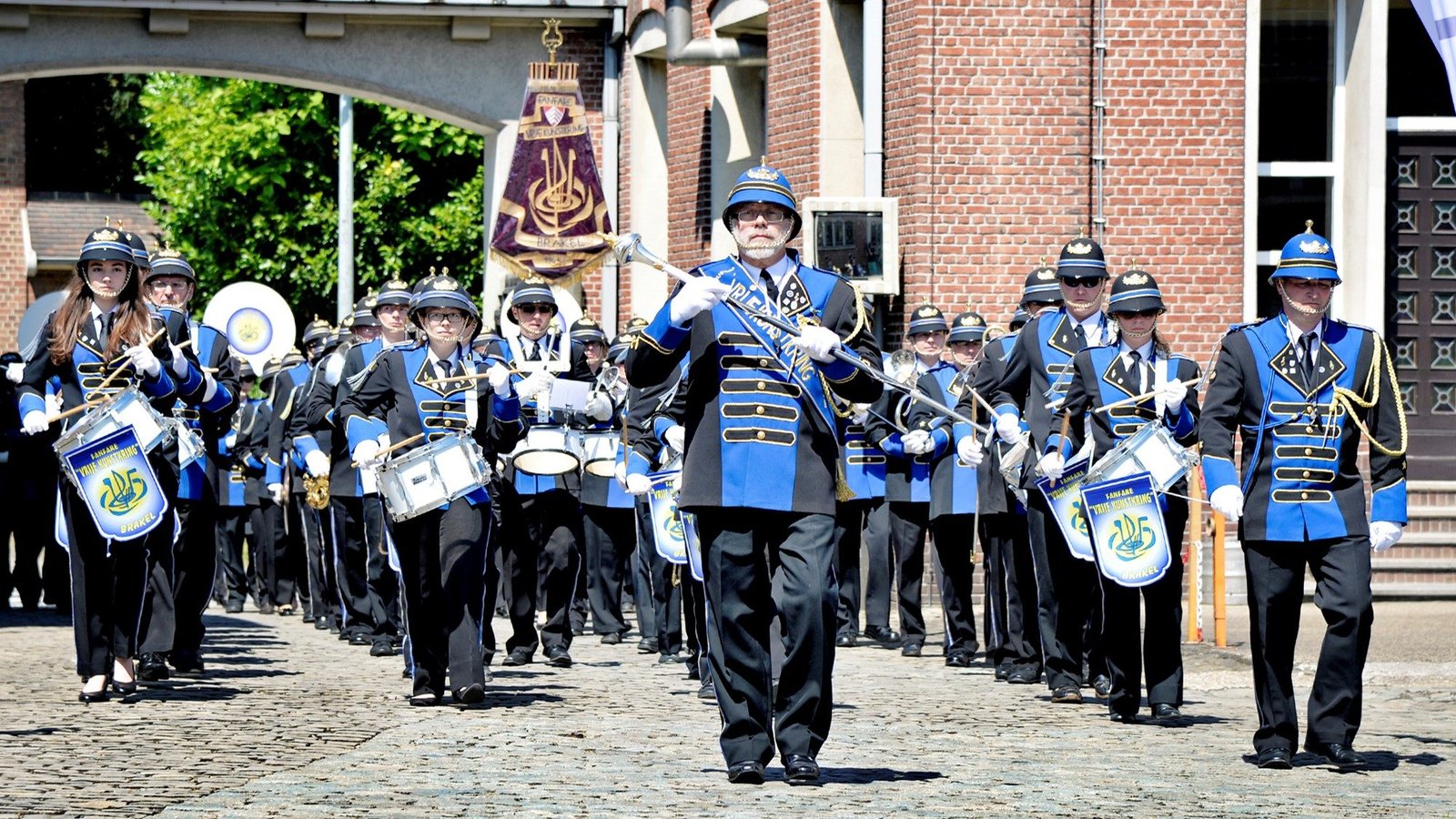 Het fanfarekorps in blauw uniform marcheert over de kasseien, met de tamboer-majoor vooraan