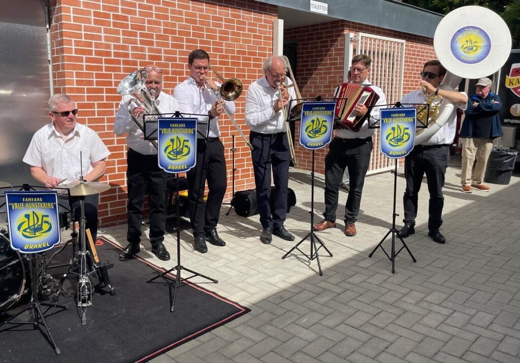 Muzikanten van Geef Moar Buzze! spelen buiten achter lessenaars met het logo van de fanfare