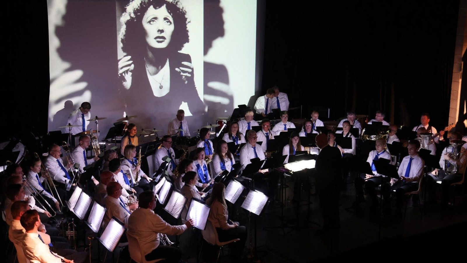 De concertband speelt onder een grote projectie van Edith Piaf